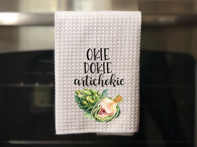 white kitchen towel with a green painted artichoke that reads okie dokie artichokie in black font, hanging on an oven door 