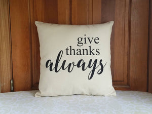 canvas pillow with black font that reads give thanks always, leaning against a wooden door 