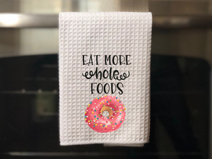 White kitchen towel is a pink donut picture that reads eat more whole foods in black writing, hanging from an oven door