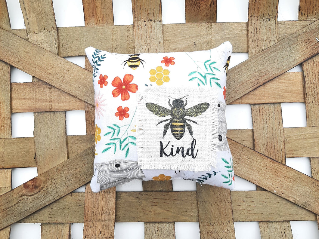 Bee kind pillow with colorful flowers and bees on it against a woven basket background 