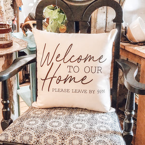 Pillow that saysWelcome to our Home Please Leave by 9pm, sitting in a decorative chair