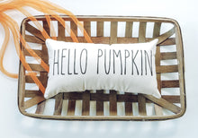 decorative canvas pillow that reads hello pumpkin in black font, sitting in a woven basket decorated with orange ribbon