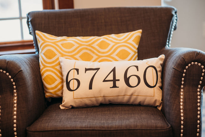 customizable zip code pillow that reads 67460 with the custom state outlined around it, sitting in an upholstered chair 