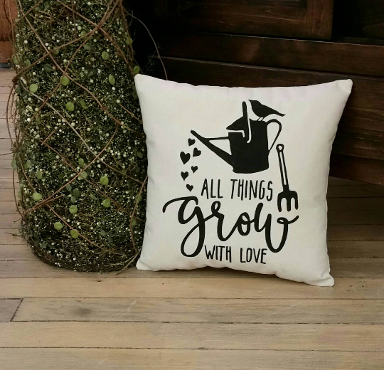 Pillow that says All Things Grow With Love with black outline of a watering can pouring out hearts leaning against a green decorative tree on a hardwood floor 