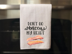 white kitchen towel with pictures of bacon that reads don't go bacon my heart in black writing, hanging on an oven door