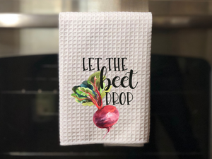 white kitchen towel with colorful beet drawn on it that reads let the beet drop in black writing, hanging on an oven door 