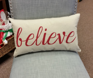 the word believe in cursive red font on a canvas pillow sitting on a grey chair