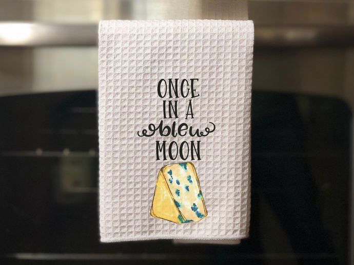white kitchen towel with a slice of bleu cheese painted on it that reads once in a bleu moon in black font, hanging on an oven door 