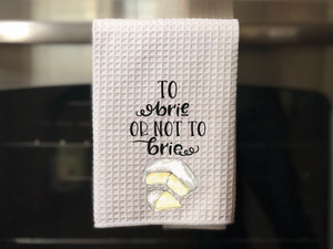 white kitchen towel with a painted piece of brie cheese that reads to brie or not to brie, hanging on an oven door