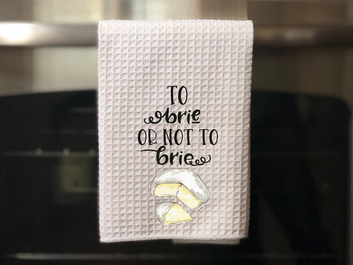white kitchen towel with a painted piece of brie cheese that reads to brie or not to brie, hanging on an oven door