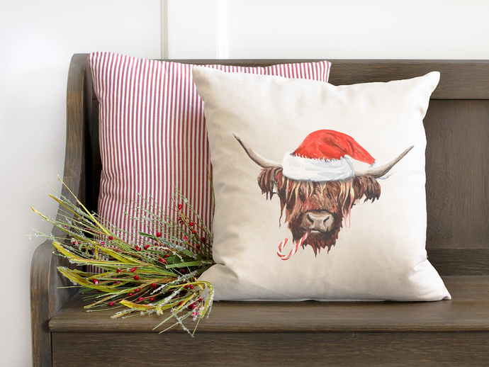 Holly Jolly Highland Cow Christmas Pillow leaning against another red and white pillow striped pillow, both sitting on a wooden bench surround by winter garland 