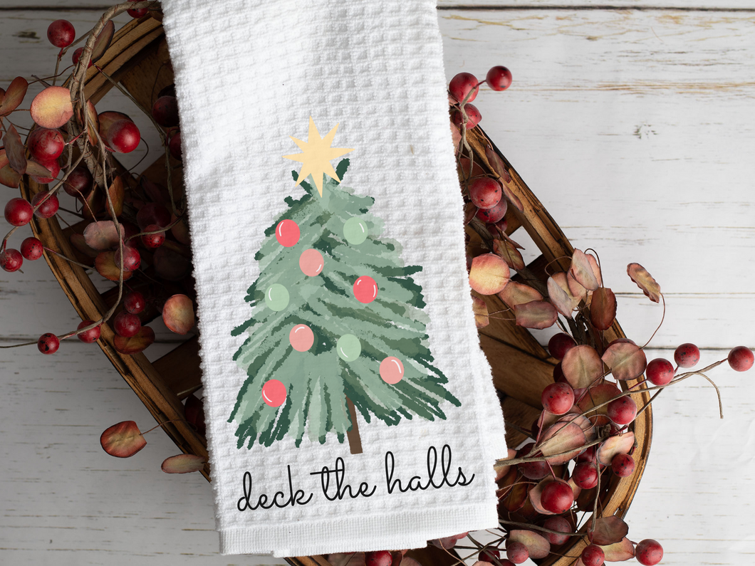 Deck the Halls Watercolor Christmas Tree Tea Towel laying across a woven basket with decorative leaves that is sitting on a white background