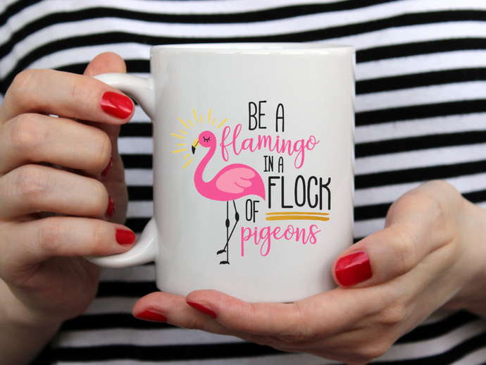 hands with red nail polish holding a mug that says Be a flamingo in a flock of pigeons against a black and white striped background