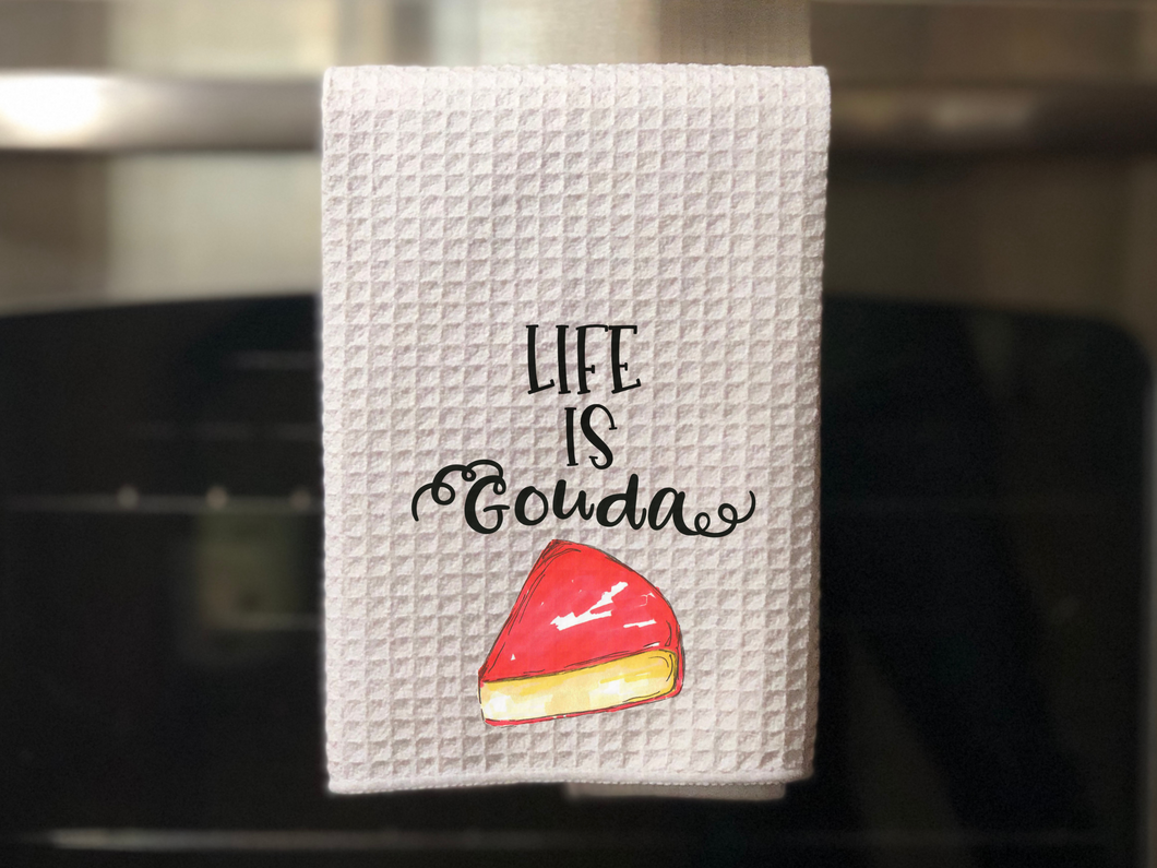 white kitchen towel with a slice of gouda cheese painted on it that reads life is gouda in black font, hanging from an oven door 