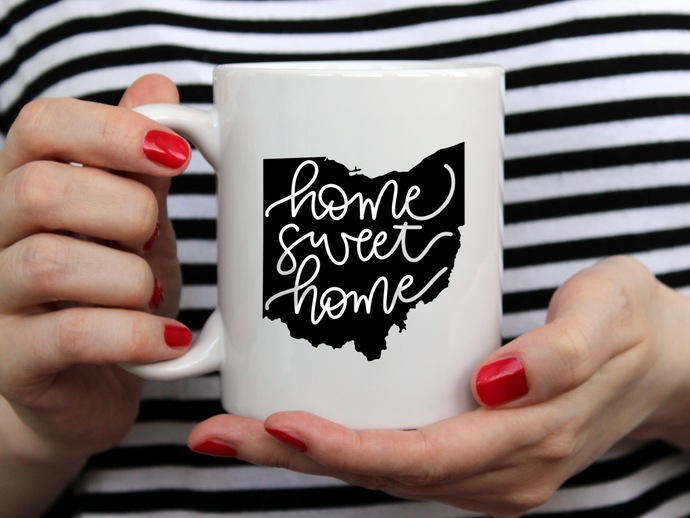 hands with red nail polish holding a white mug with a black outlined state of Ohio that reads home sweet home
