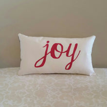 Joy Christmas pillow leaning against a tan wall