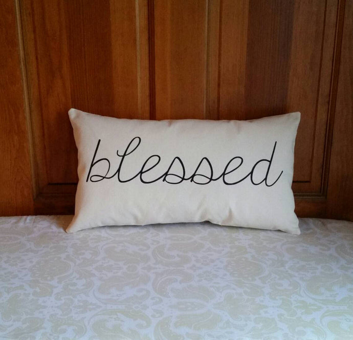 pillow that reads blessed in black cursive font, leaning against a wooden wall