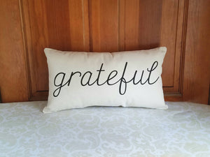 modern farmhouse pillow that reads grateful in black cursive font, leaning against a wooden wall