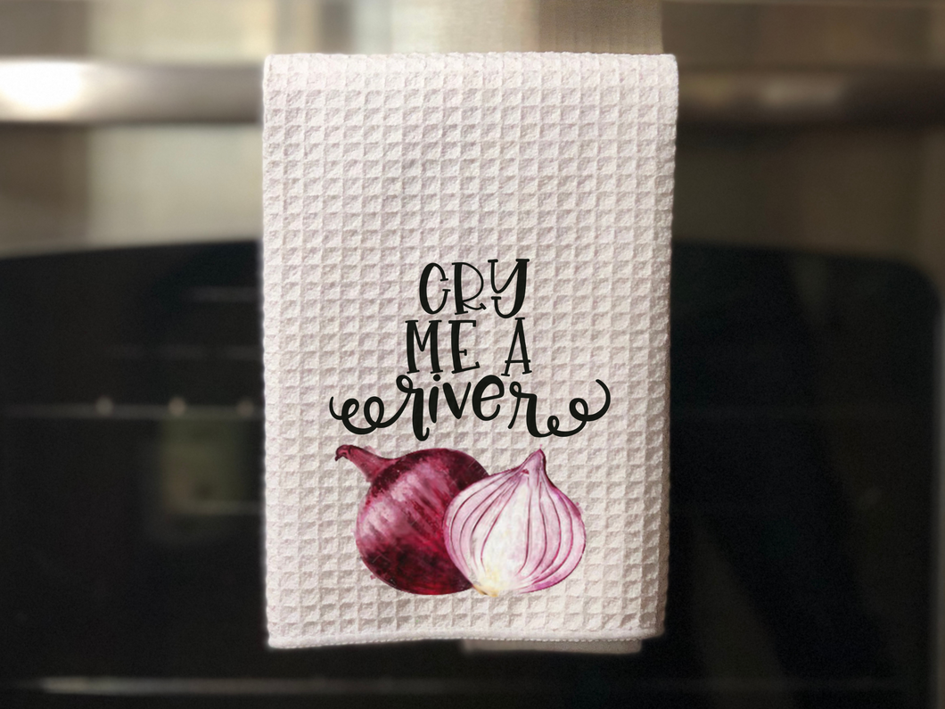 white kitchen towel with a purple onion on it that read cry me a river in black writing, hanging on an oven door