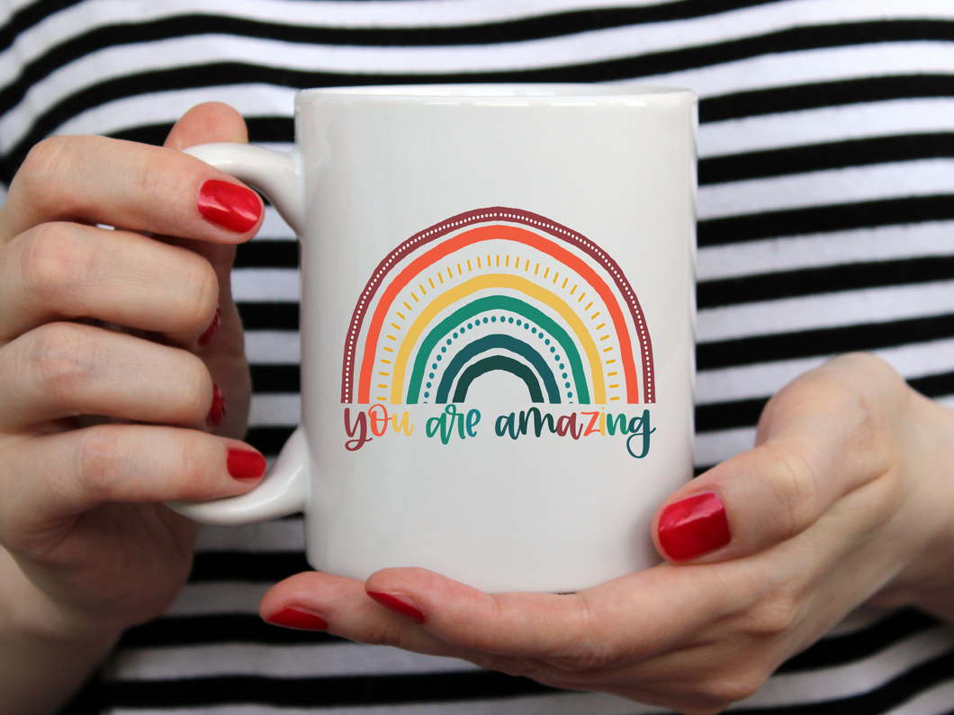 hands with red nail polish holding a white mug with a colorful rainbow on it and you are amazing in colorful font 